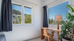 Une pièce avec un bureau en bois, une chaise, une lampe, une plante en pot et une vue sur un immeuble et des arbres, visible à travers les fenêtres.