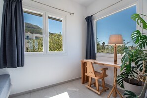 Une pièce avec un bureau en bois, une chaise, une lampe, une plante en pot et une vue sur un immeuble et des arbres, visible à travers les fenêtres.