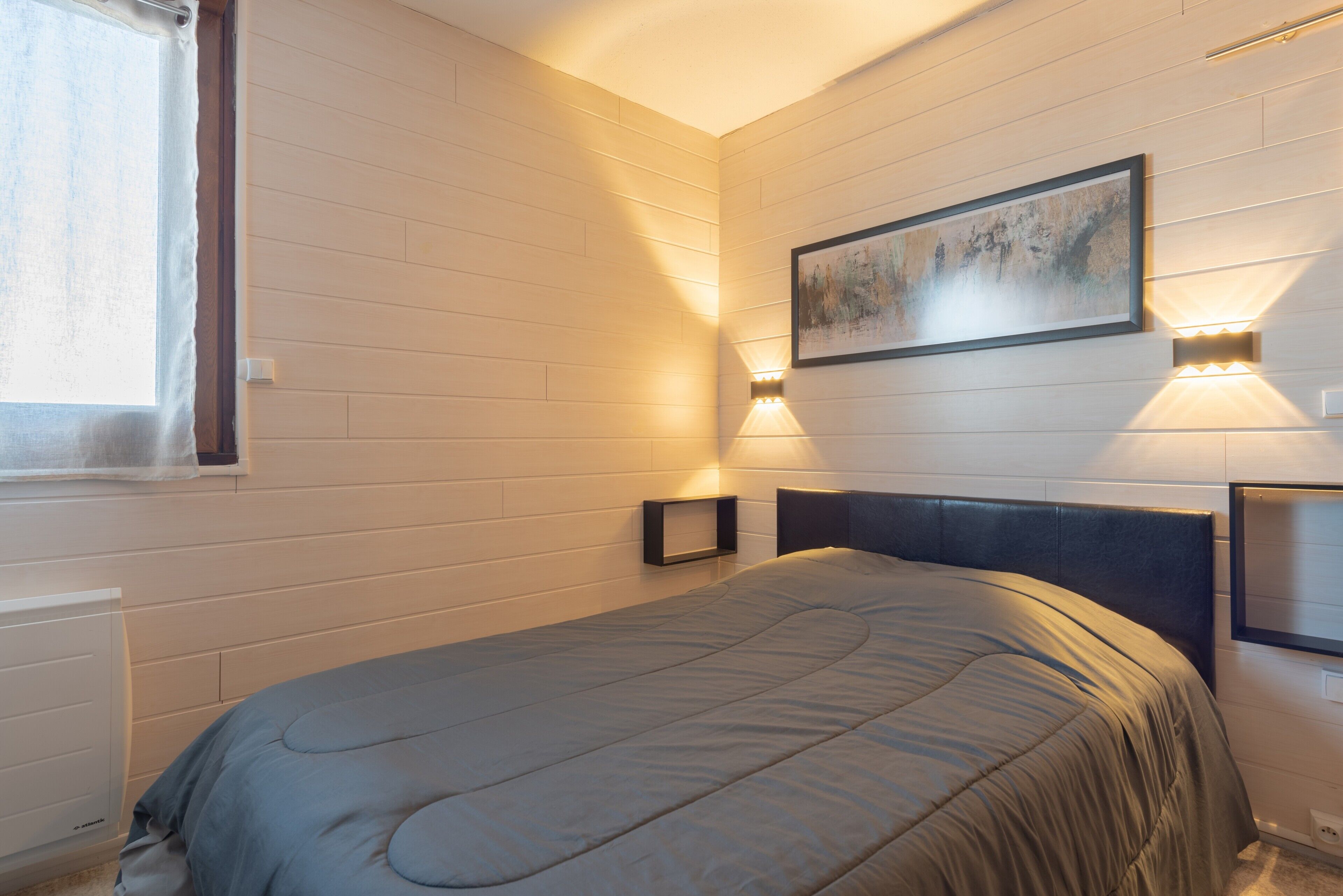 Un dormitorio con cama, ventana e iluminación en la pared.