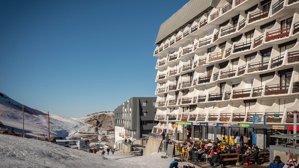 Un hotel de varios pisos con balcones, una pista de esquí al fondo y zonas de estar al aire libre.