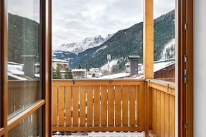 Un balcone con ringhiera in legno che si affaccia su un paesaggio montuoso con cime innevate e edifici.