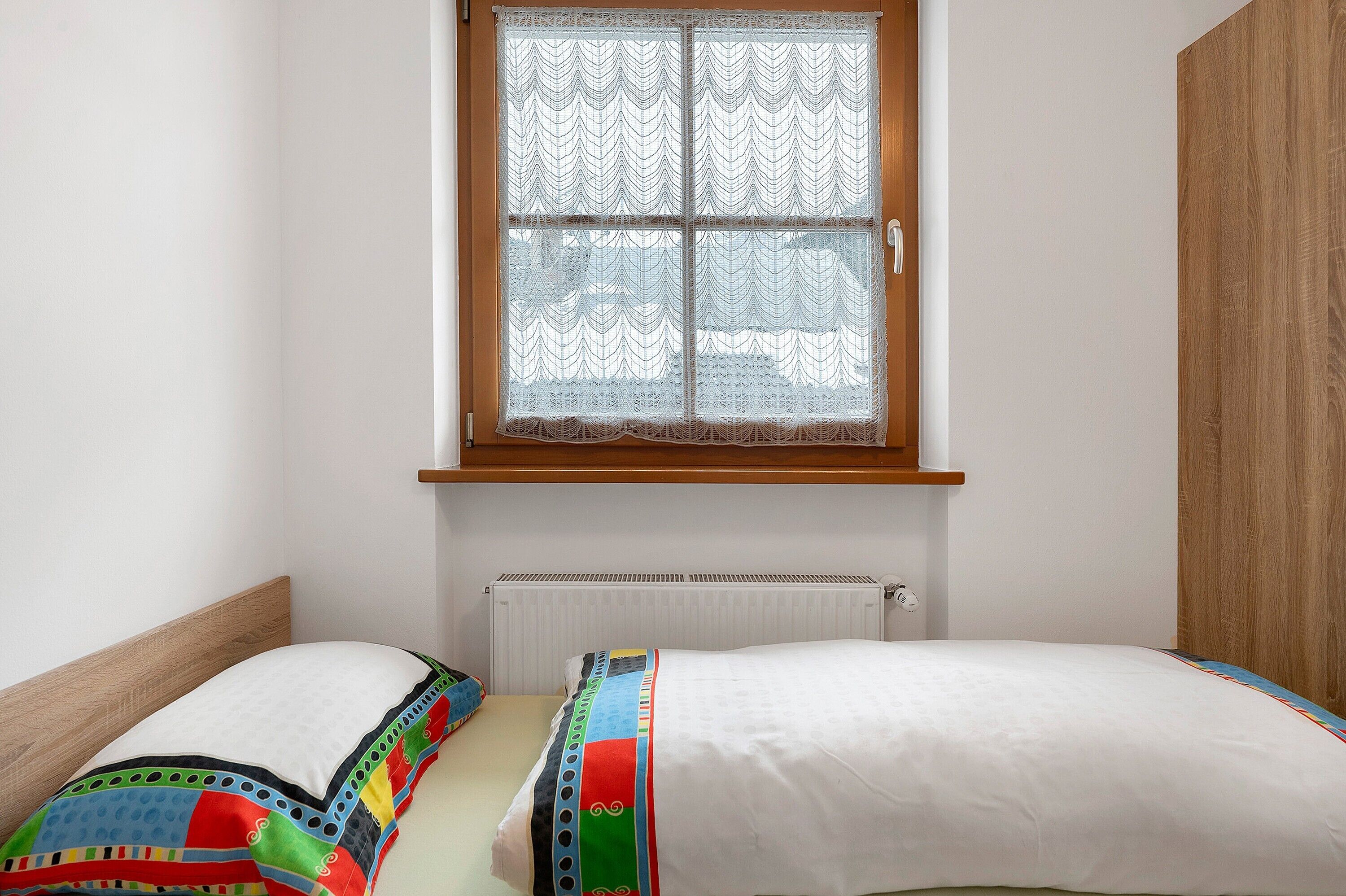 A small room with a window, a radiator, and a bed with a colorful bedspread.