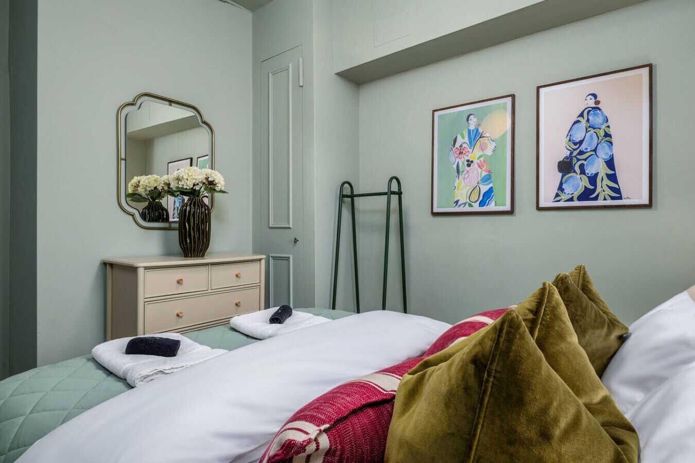 Ein ordentlich eingerichtetes Schlafzimmer mit einem Bett, einem Schminktisch, einem Spiegel und zwei gerahmten Kunstwerken an der Wand.