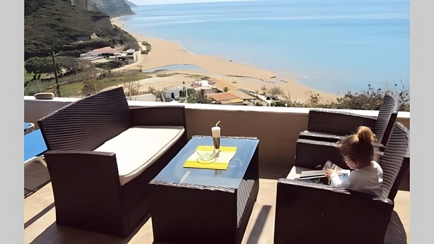 A balcony with a view of the beach, a table with a candle, and a person reading.