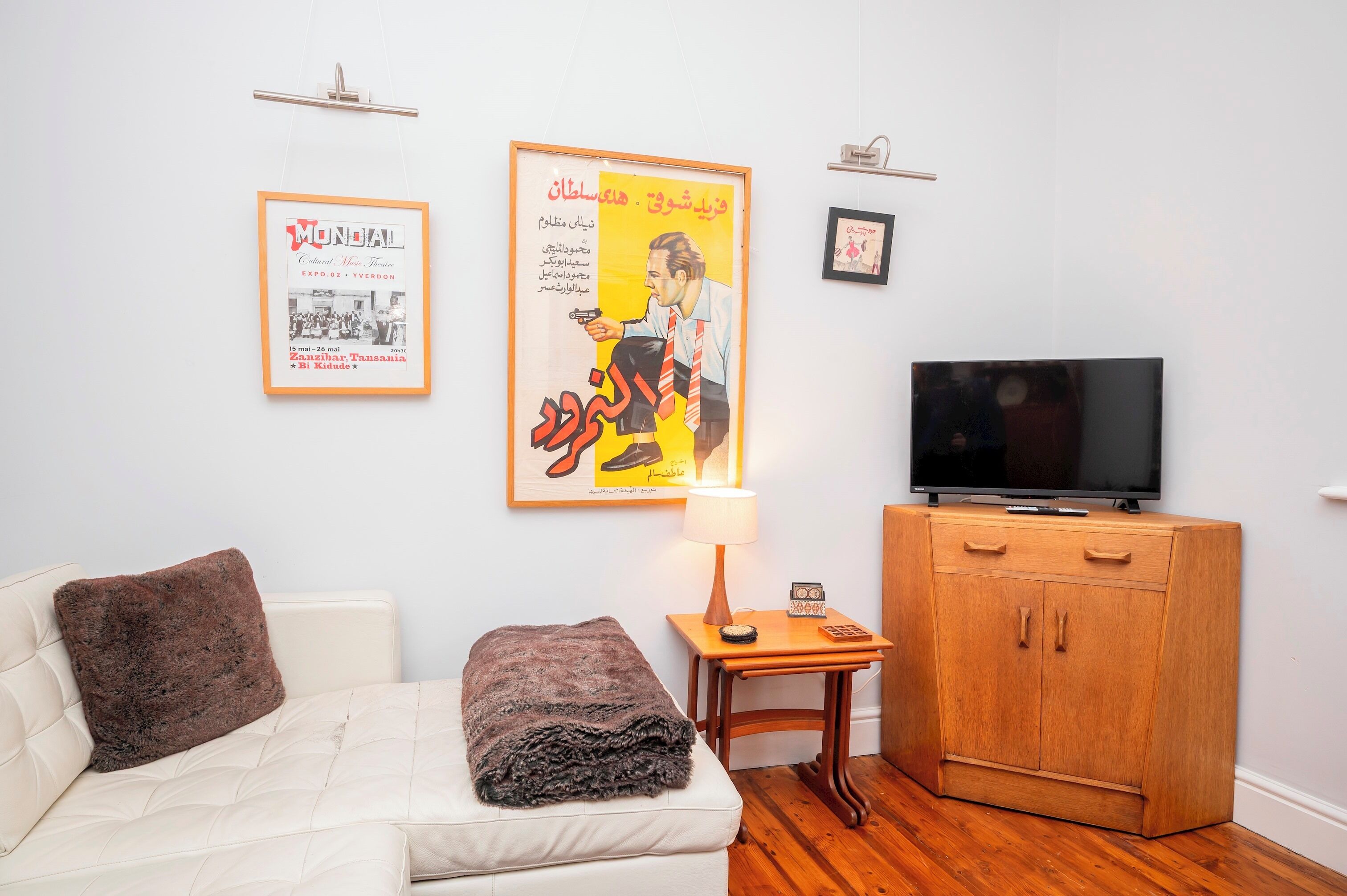 A living room with a white sofa, a brown throw blanket, a wooden cabinet, a TV, and two framed posters on the wall.