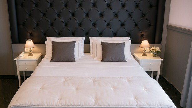 Una habitación de hotel con una cama grande, dos mesitas de noche, un cabecero tapizado negro y una mesita con una lámpara y flores.