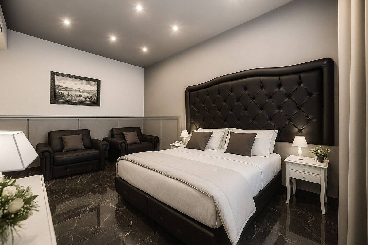 A modern hotel bedroom with a large bed, two armchairs, a nightstand, and a framed picture on the wall.