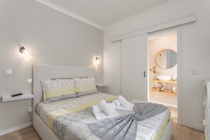 A bedroom with a bed, bedside tables, a nightstand, a wall-mounted light, a mirror, and a bathroom with a sink and towel rack. - Holiday Home 'Refúgio das Barcas' with Sea View, Wi-Fi & Air Conditioning (Lourinha)