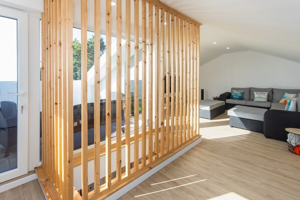 Un salon moderne avec une cloison en bois, un canapé gris et une porte vitrée donnant sur l’extérieur.