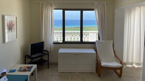 A room with a TV, a white chair, a small table, and a window with a view of the sea. - NiceView Apartment in Son Bou with Sea View – Ideal for Couples (ALAIOR)