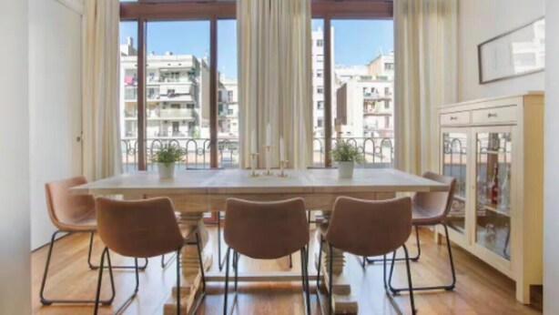 Ein Essbereich mit einem großen Tisch, vier Stühlen, einem Fenster mit Vorhängen und Blick auf Gebäude.