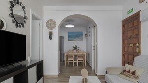 Une chambre d’hôtel équipée d’un téléviseur à écran plat, d’un canapé agrémenté de coussins à motifs, d’une table à manger en bois avec des chaises, et d’un tableau représentant une scène de plage.