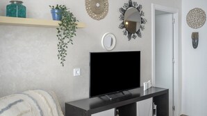 Un salon avec un téléviseur à écran plat, une plante, des éléments décoratifs suspendus au mur et une étagère sur laquelle se trouvent un bocal et une plante en pot.