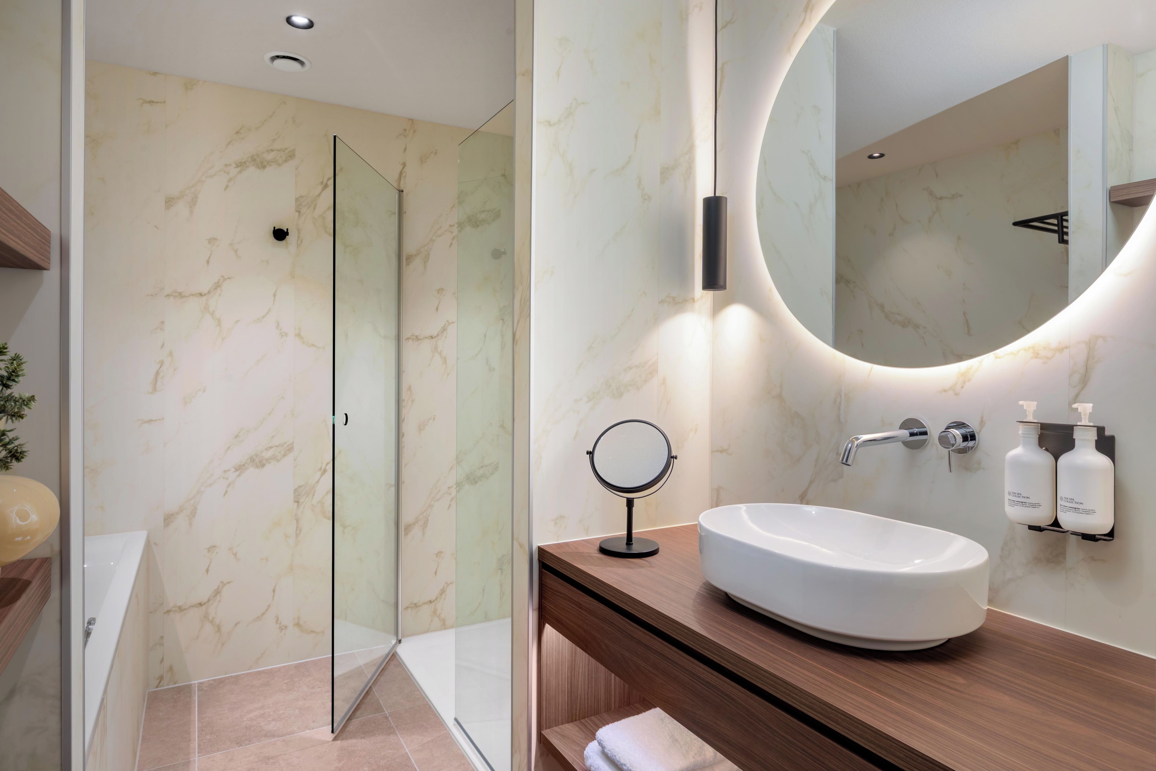 A modern bathroom with a glass shower enclosure, a round mirror, a white sink, and two soap dispensers.