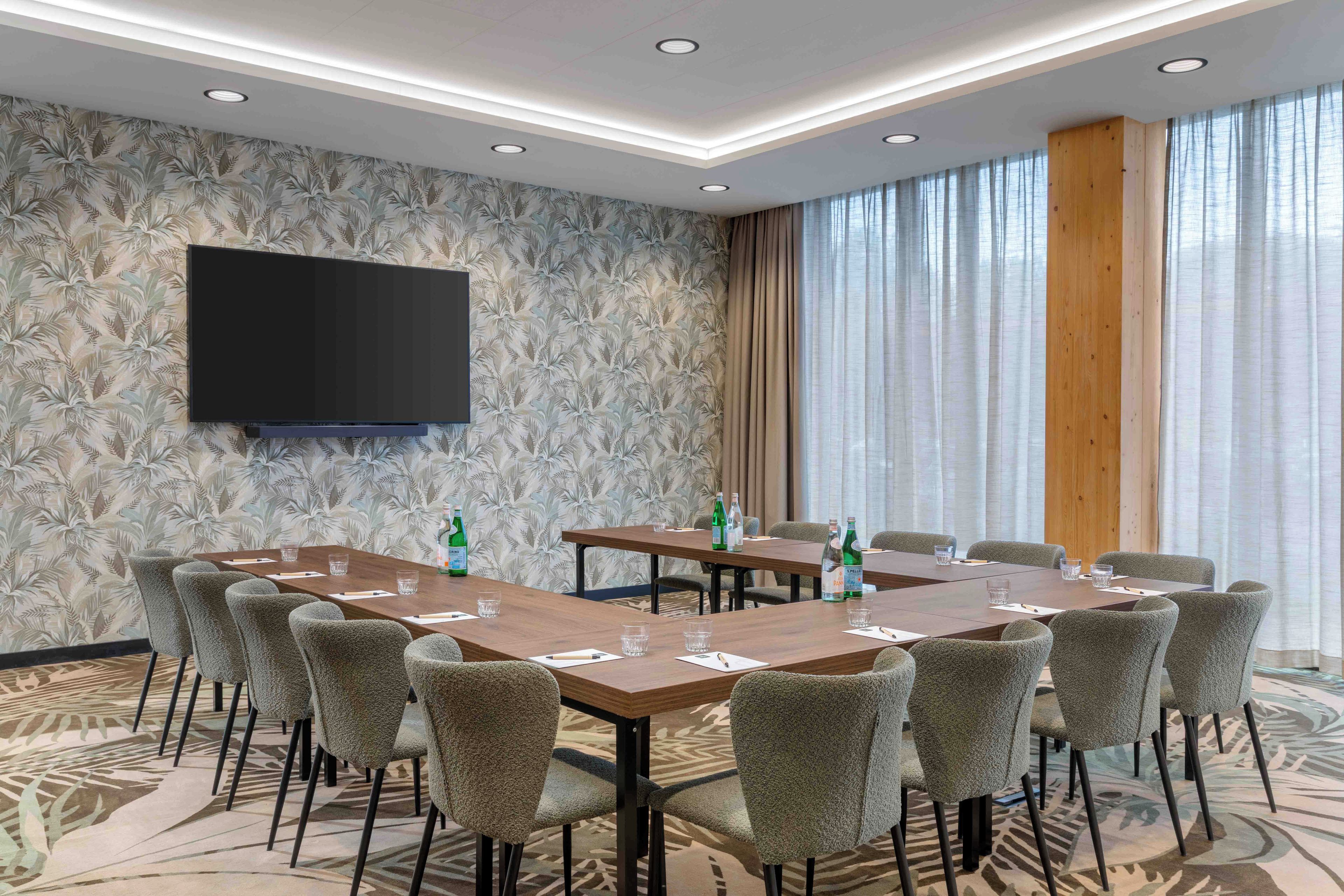 A conference room with a large flat-screen TV mounted on the wall, multiple tables with chairs, and a patterned carpet.