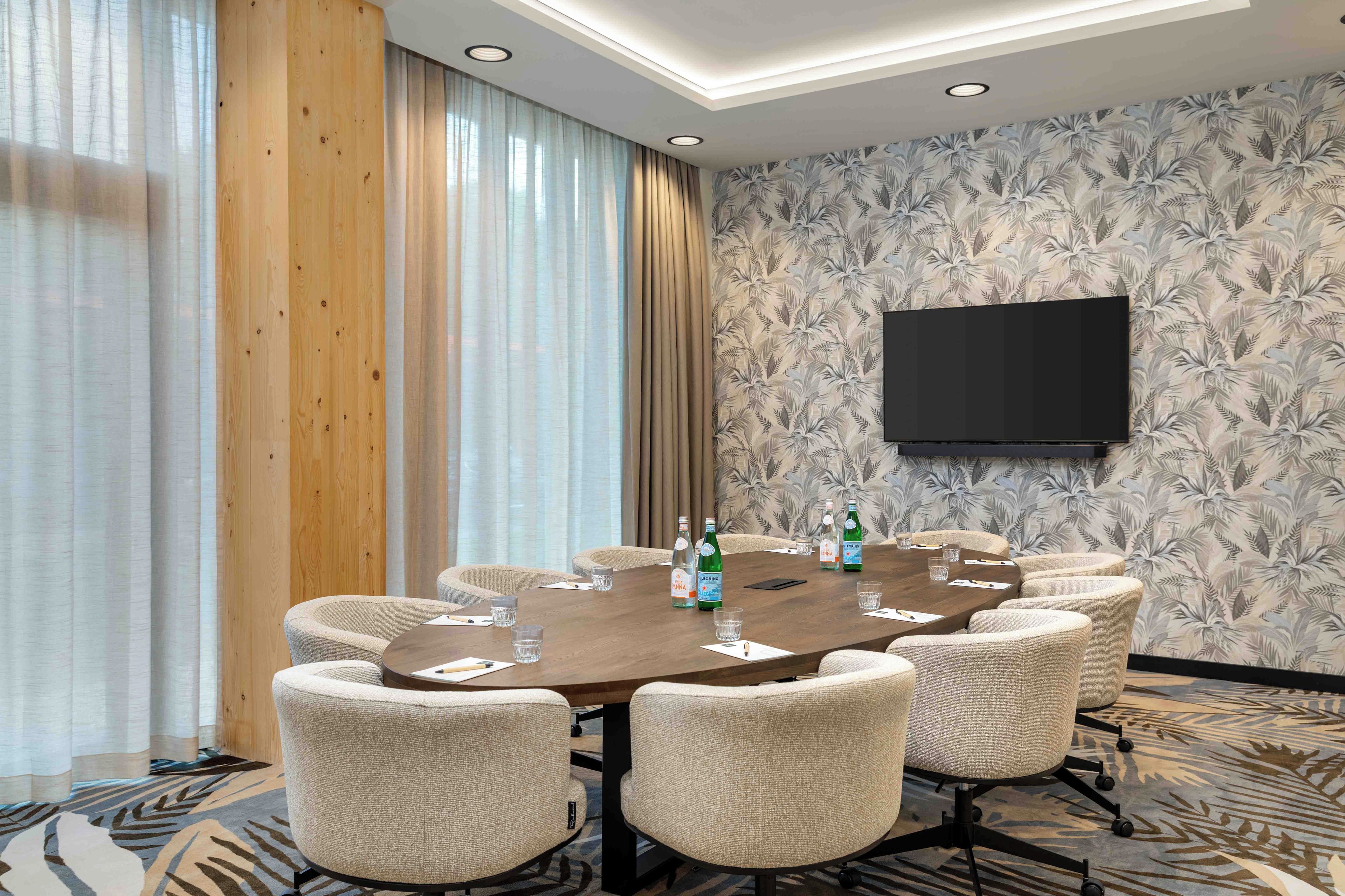 Une salle de conférence avec une table ronde, six chaises, un téléviseur à écran plat fixé au mur et un papier peint à motifs.