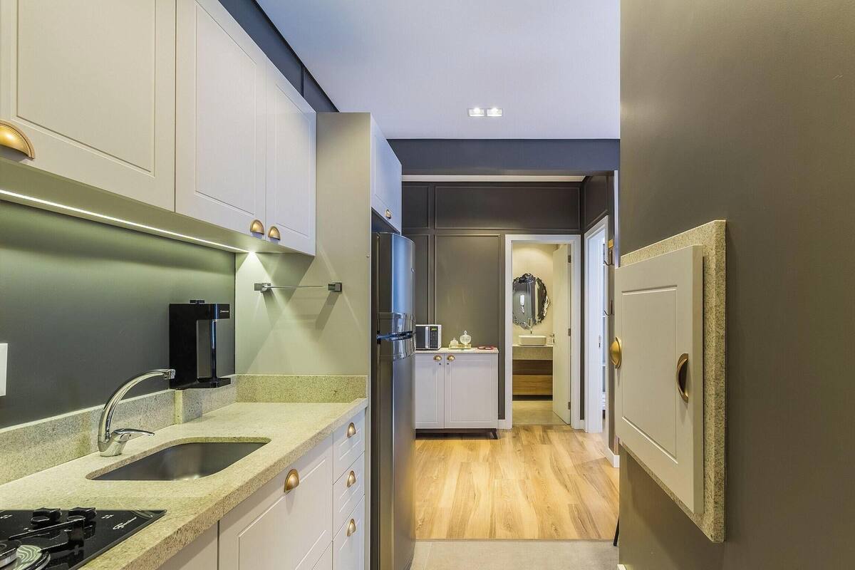 Un couloir étroit, avec un coin cuisine sur la gauche, comprenant un évier, une cuisinière et des placards. Une porte donne accès à une salle de bain équipée d’un miroir et d’un évier.