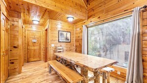 A wooden cabin interior with a large dining table, benches, a framed picture on the wall, and a large window with curtains. - 4BR Cabin for 8 w/ Game Room & Hot Tub Haven! (Sevierville)