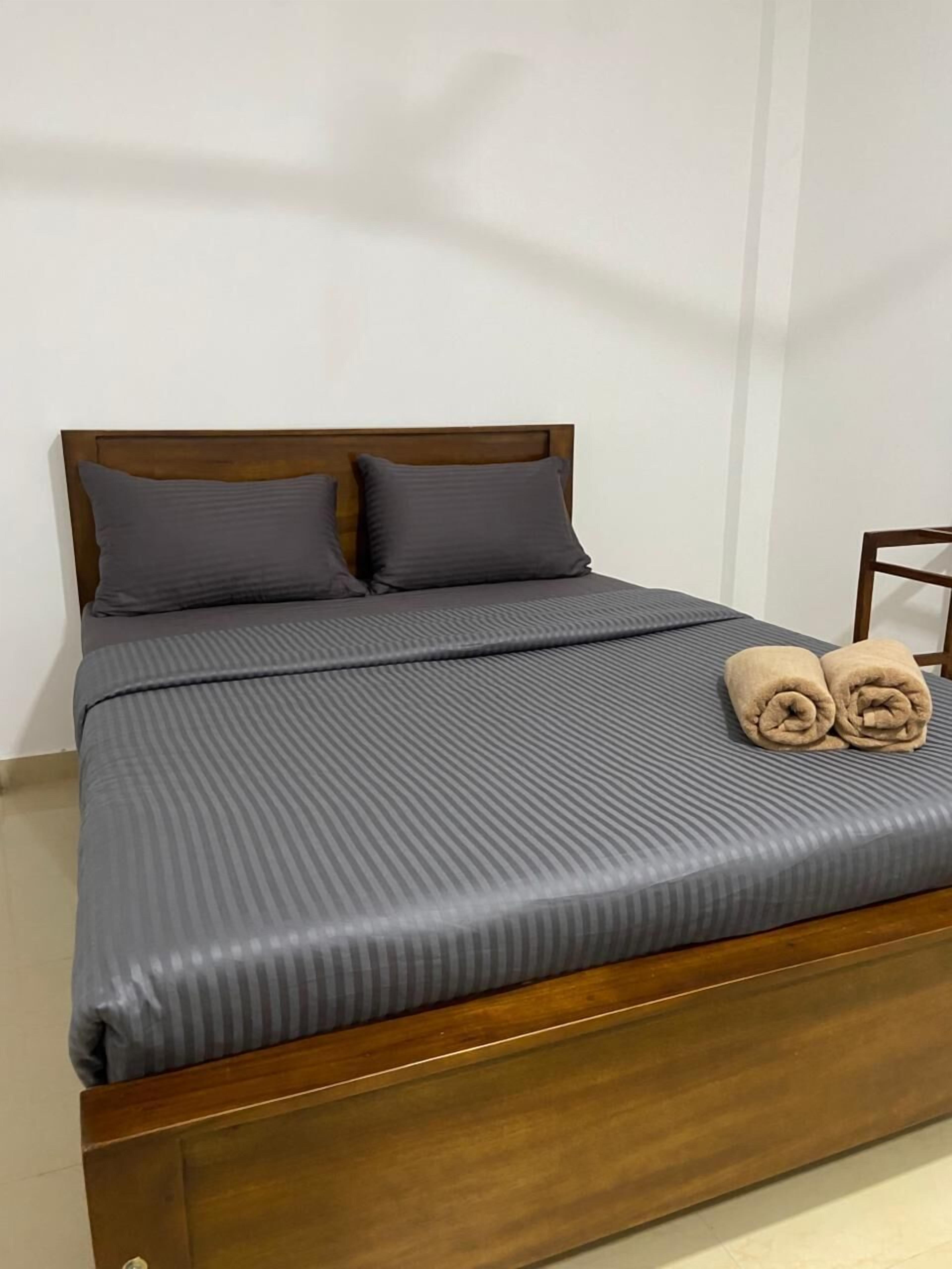 A neatly made bed with a wooden headboard, grey bedding, and two rolled towels on the footboard.