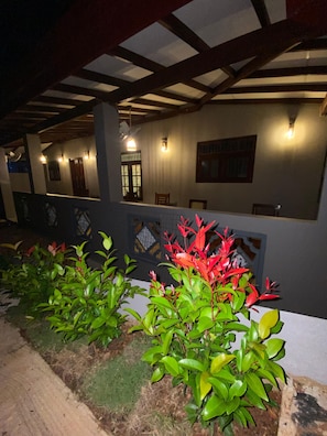 Un espacio exterior con una zona de estar cubierta, plantas y un muro con ventanas y puertas.