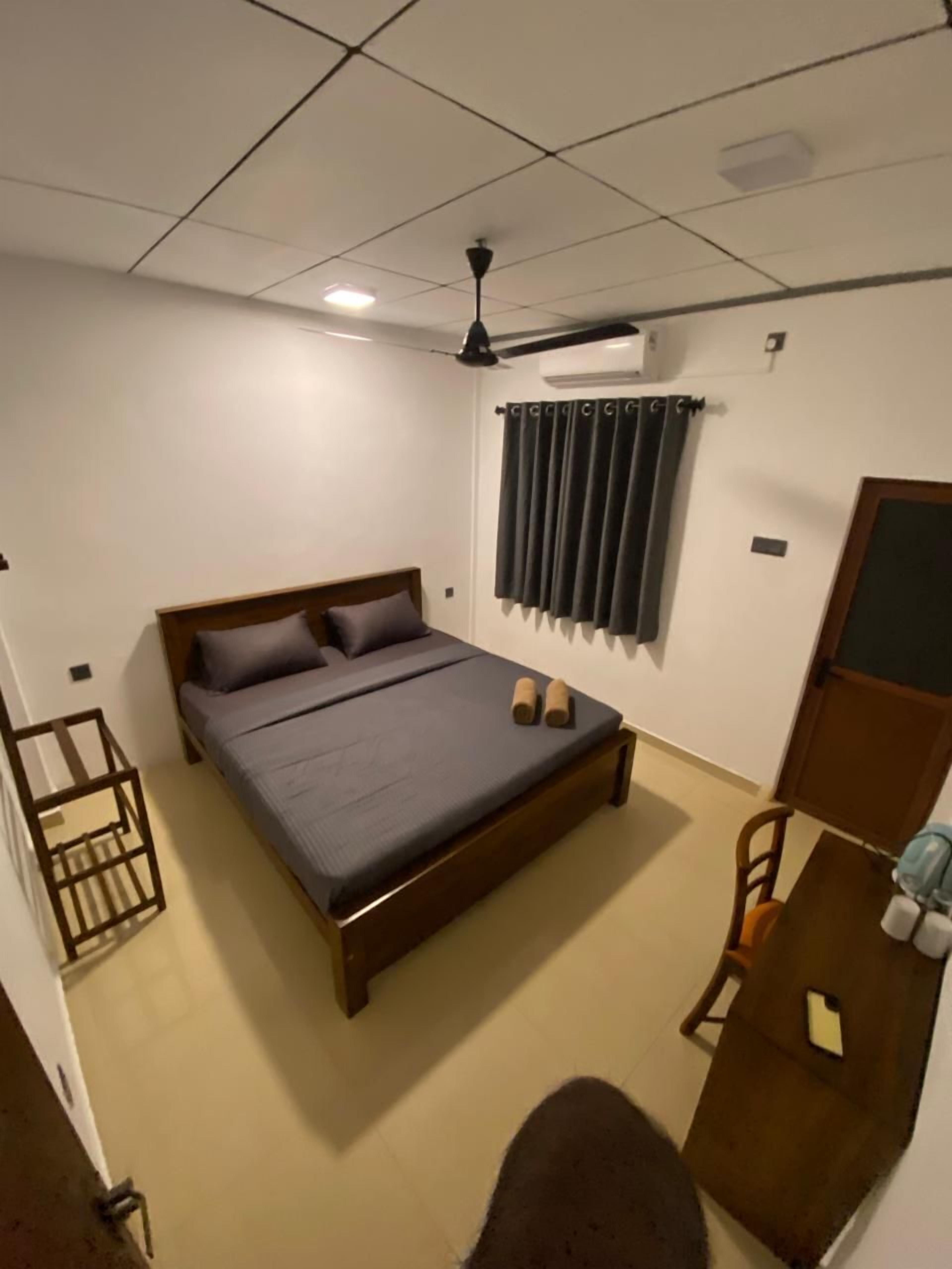 A room with a bed, a table, a chair, a ceiling fan, and a window with curtains.