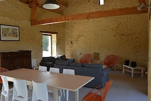 Ein rustikales Interieur mit Steinwand, Holzbalken, einer Couch, einem Esstisch mit Stühlen und einem roten Sessel.