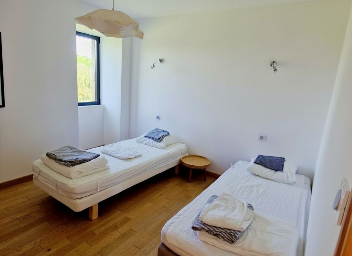 A room with two single beds, a small round table, a window with a view, and a hanging light fixture.