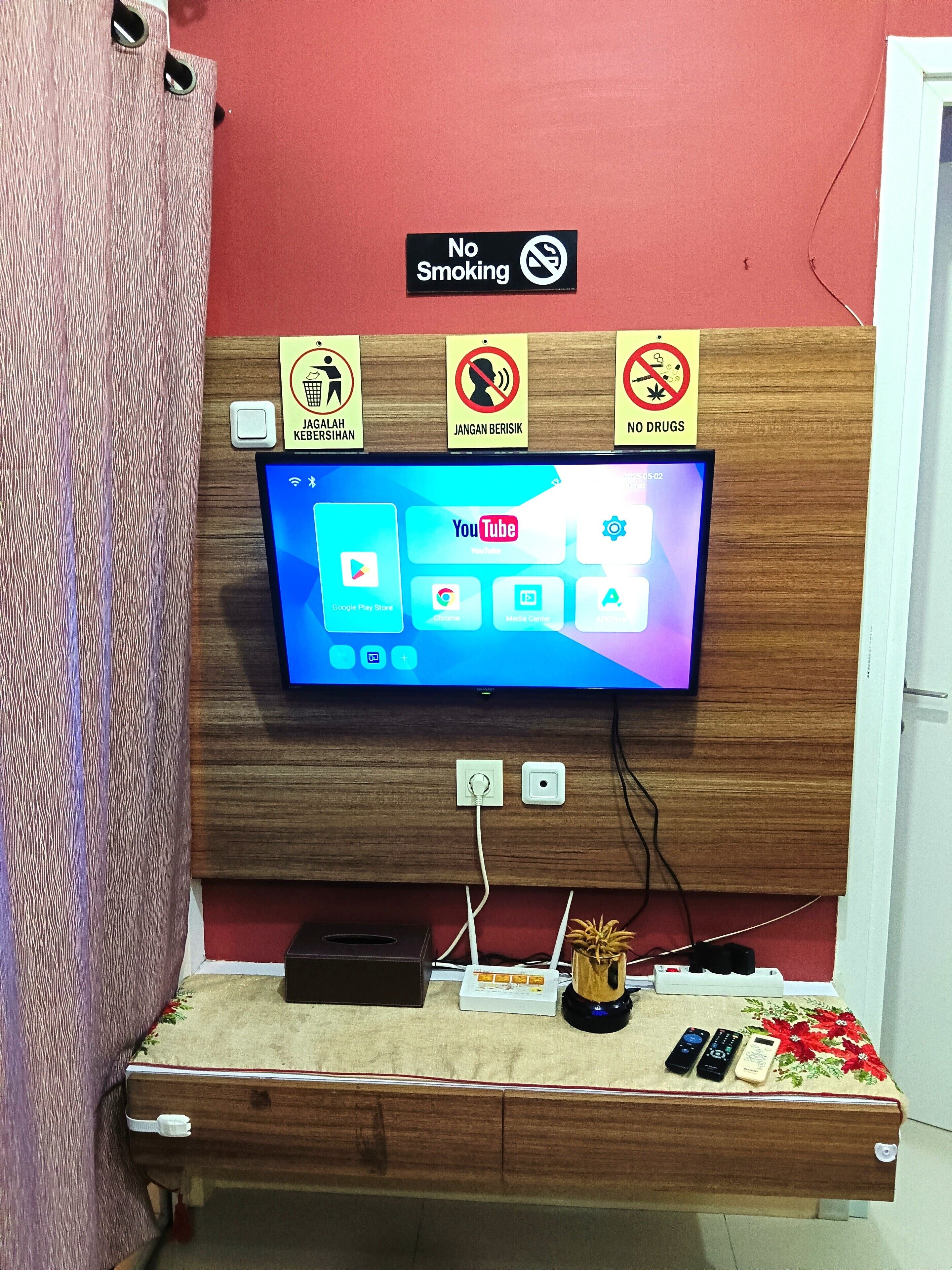 A hotel room with a mounted television displaying various apps, a wooden headboard, and a sign indicating no smoking, no drugs, and no littering.