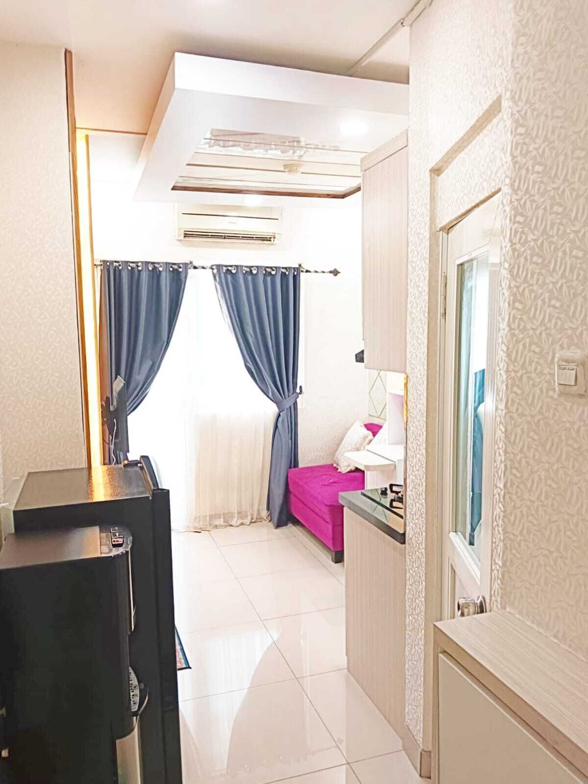 A compact hotel room with a kitchenette, a bed with a pink bedspread, a desk with a lamp, and a window with patterned curtains.