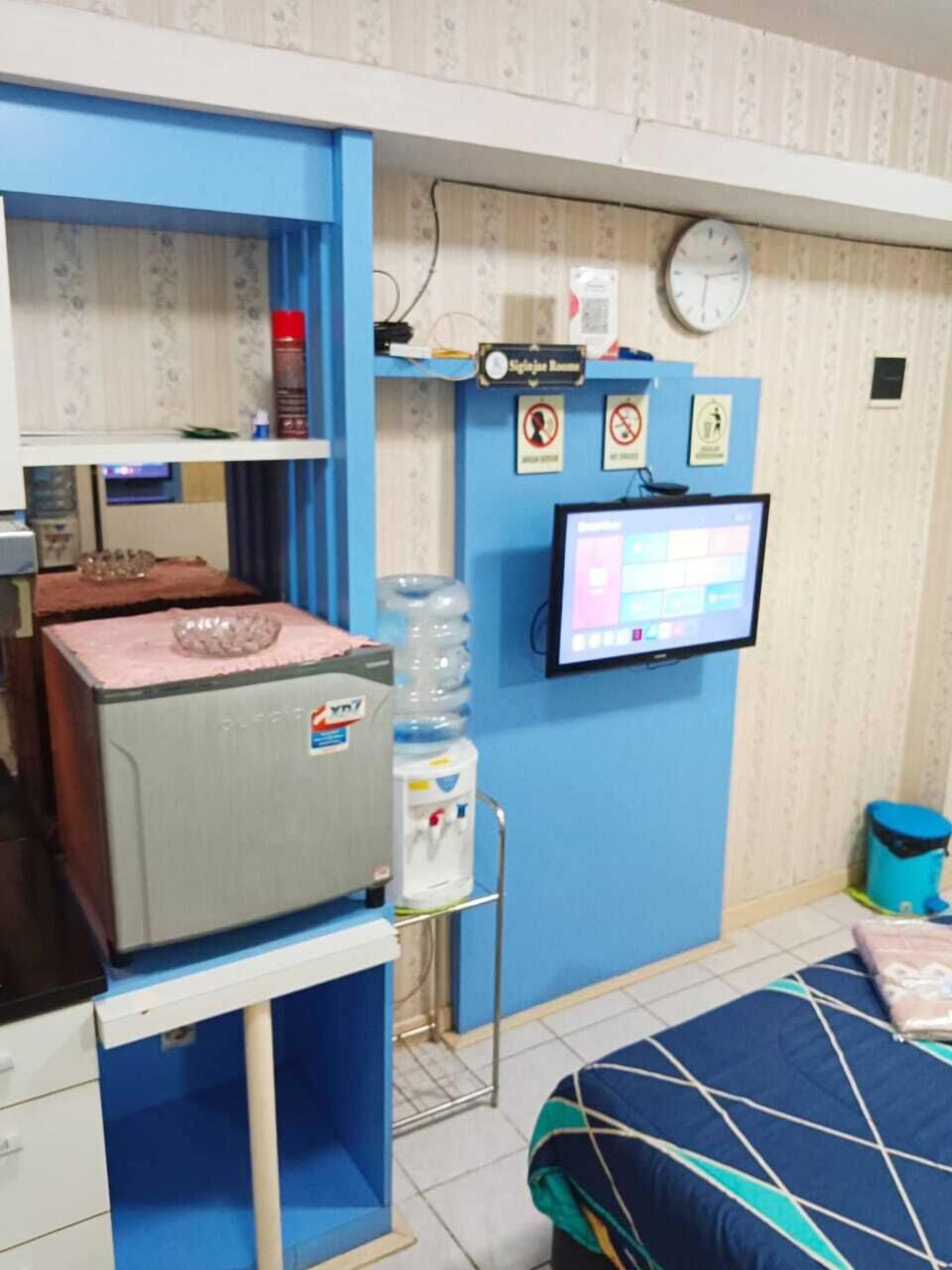 Een kamer met een bed, een bureau met een computer, een waterdispenser en een klok aan de muur.