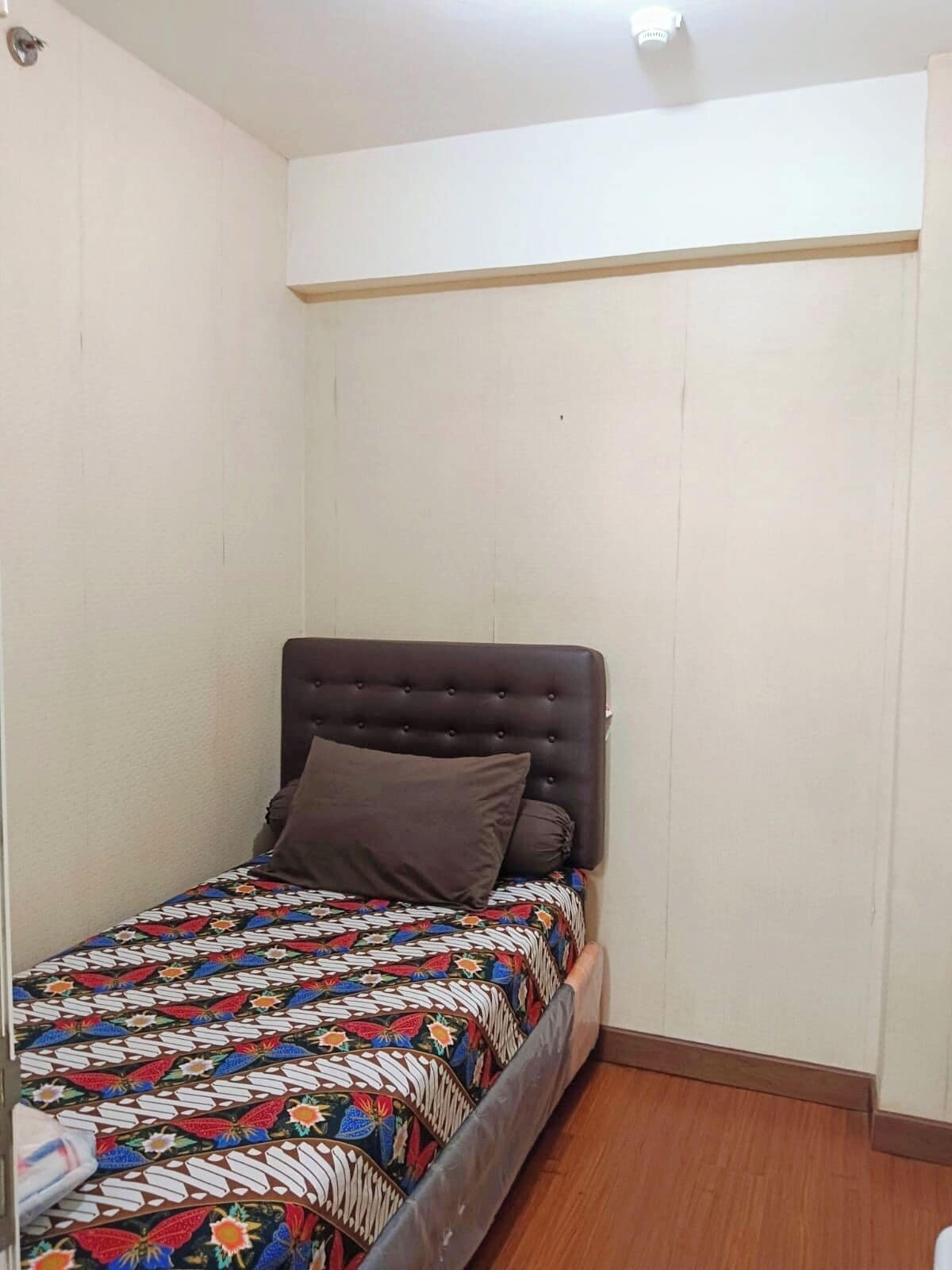 A small, single-bed room with a wooden floor, a dark headboard, and a colorful bedspread.