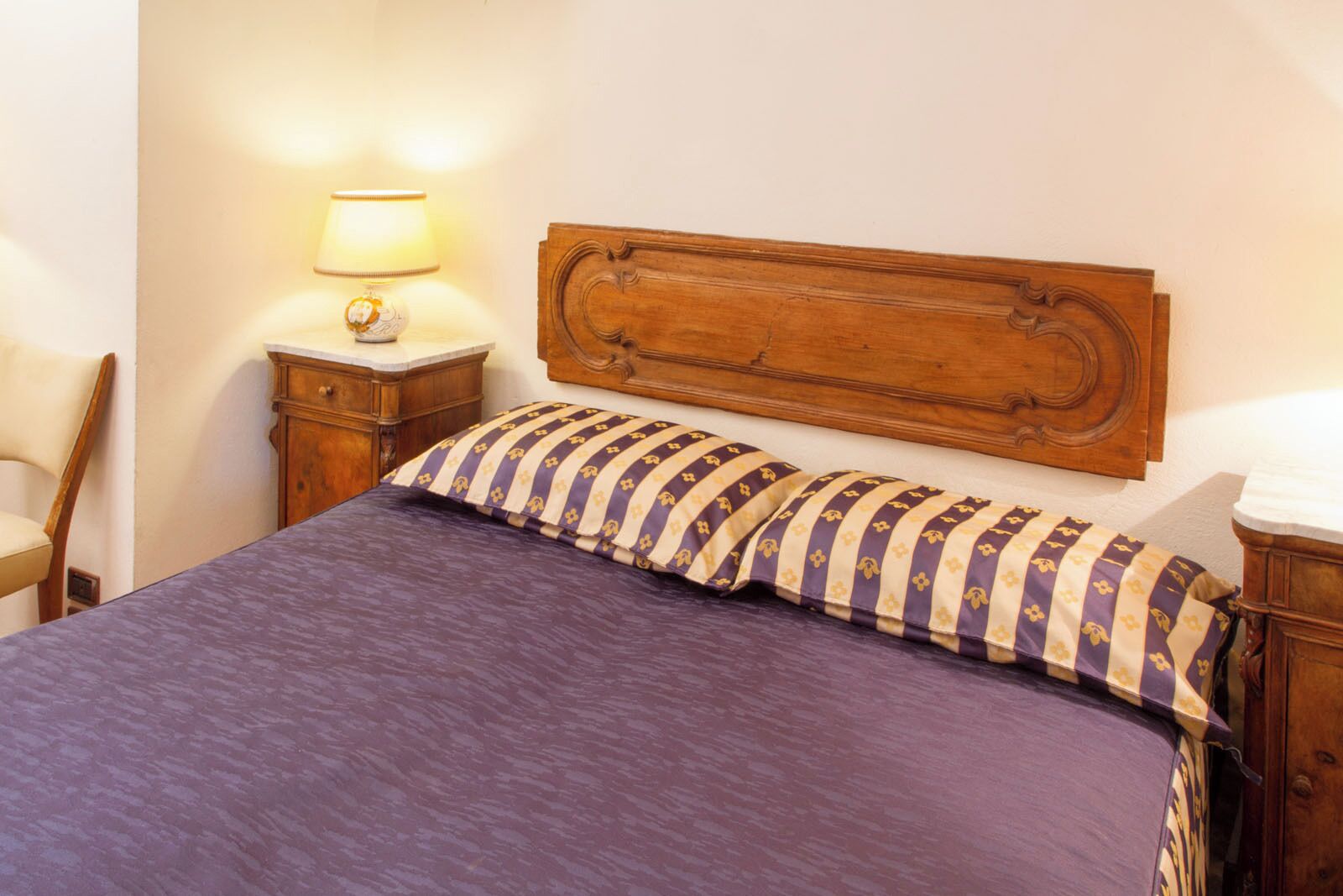 Una cama con colcha morada y almohadas a rayas, cabecera de madera, mesita de noche con lámpara y una silla de madera.