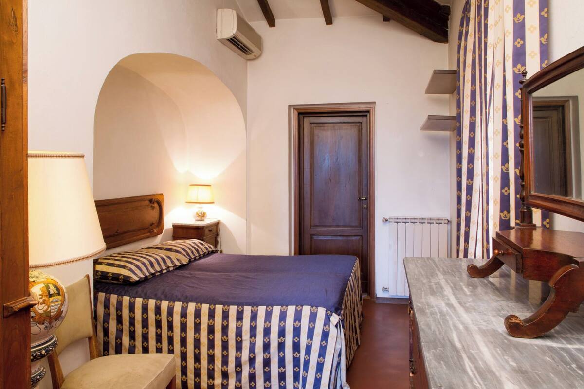 A bedroom with a bed, a nightstand, a dresser, a mirror, a door, and an arched window.