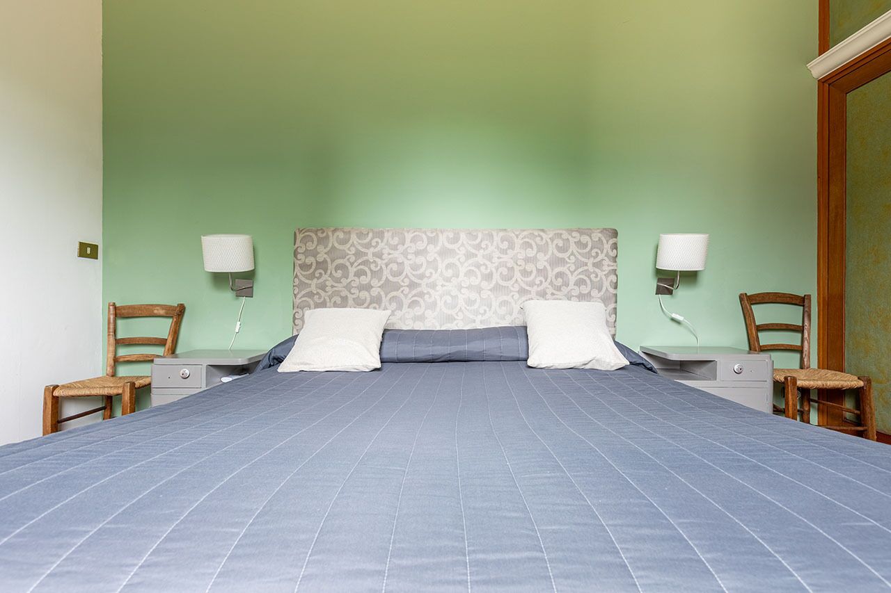 Ein ordentlich bezogenes Bett mit blauer Bettdecke und zwei Kissen, eingerahmt von zwei Nachttischen mit Lampen, in einem Zimmer mit grünen Wänden und einem gemusterten Kopfteil.