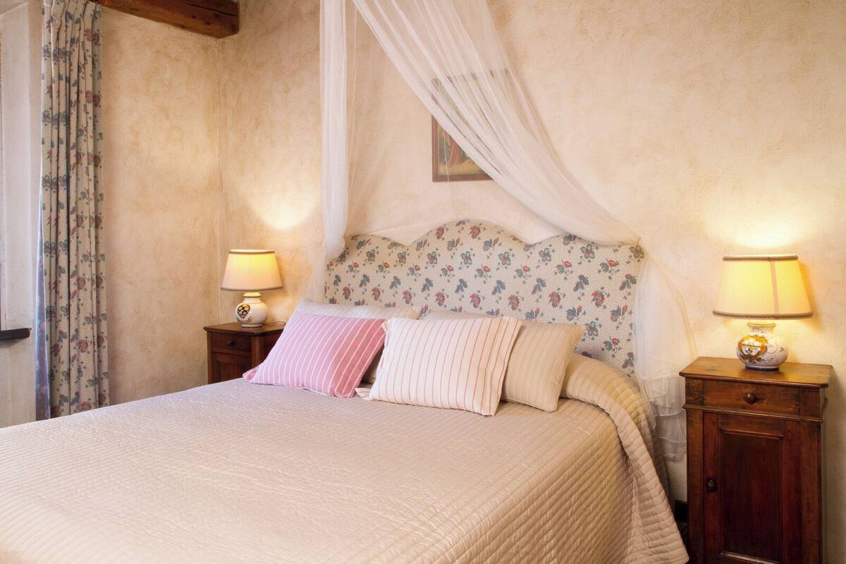 A bed with a canopy, two bedside tables with lamps, floral wallpaper, and a window with curtains.