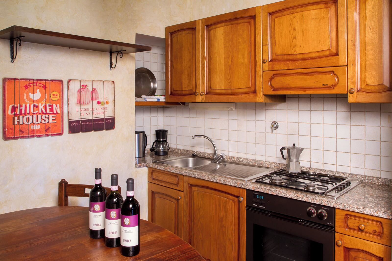Uma cozinha com armários de madeira, bancada de granito, fogão, pia e cafeteira. Há três garrafas de vinho sobre a mesa.