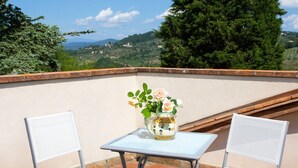 A terrace with two white chairs and a small table, a vase with flowers, and a scenic view of hills and trees. - Relais de Ricci (Impruneta)
