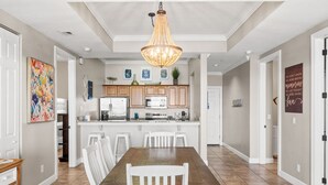 A spacious dining area with a wooden table and white chairs, a kitchen with wooden cabinets and modern appliances, and a decorative chandelier. - Huge Balcony-Spectacular Views-Penthouse 4BR/4Bath (Panama City Beach)
