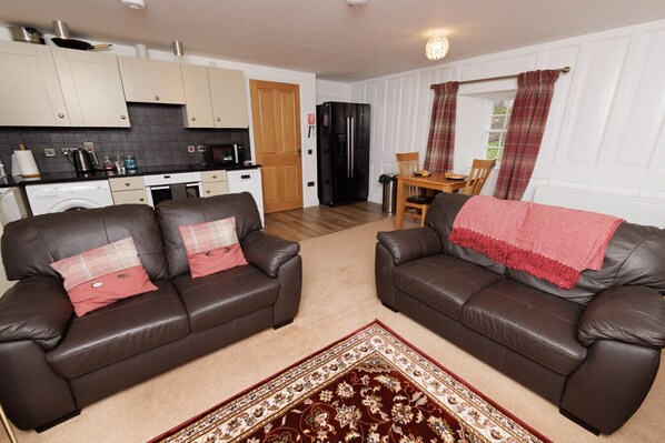 A living room with two brown leather sofas, a patterned rug, and a kitchen in the background. - NEW to VRBO: Scalloway Charm • 1BR Apt with Coastal Views (Shetland Islands)