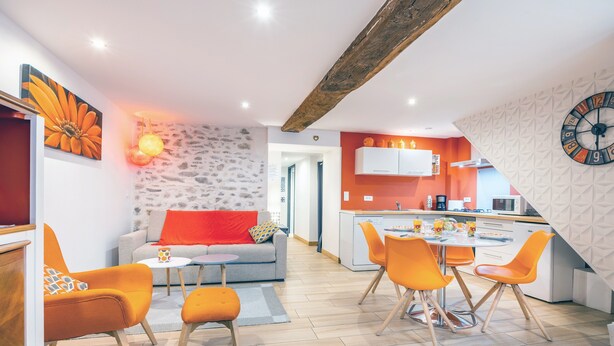 Een moderne keuken met een klok, een eethoek met oranje stoelen, en een woonkamer met een bank en een rood kussen.