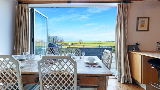 Comedor con mesa preparada para cuatro, techo de madera y vista a un campo verde a través de amplias ventanas.