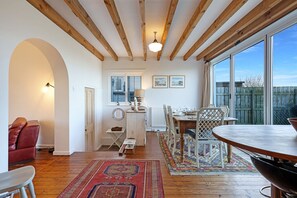 A cozy dining area with a wooden table and chairs, a red patterned rug, and a view of the outdoors through large windows. - Grove Cottage -North East Escapes (Beadnell)