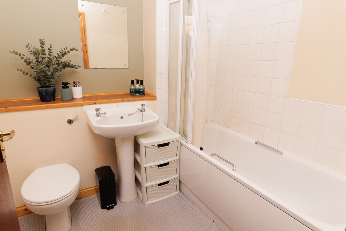 Un bagno con toilette, lavabo e vasca. Sopra il lavabo c'è uno specchio, una pianta in vaso e articoli da toilette sul piano del lavabo.