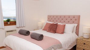 Een net opgemaakt bed met een roze hoofdbord, twee bedlampjes en uitzicht op een waterpartij door een raam.