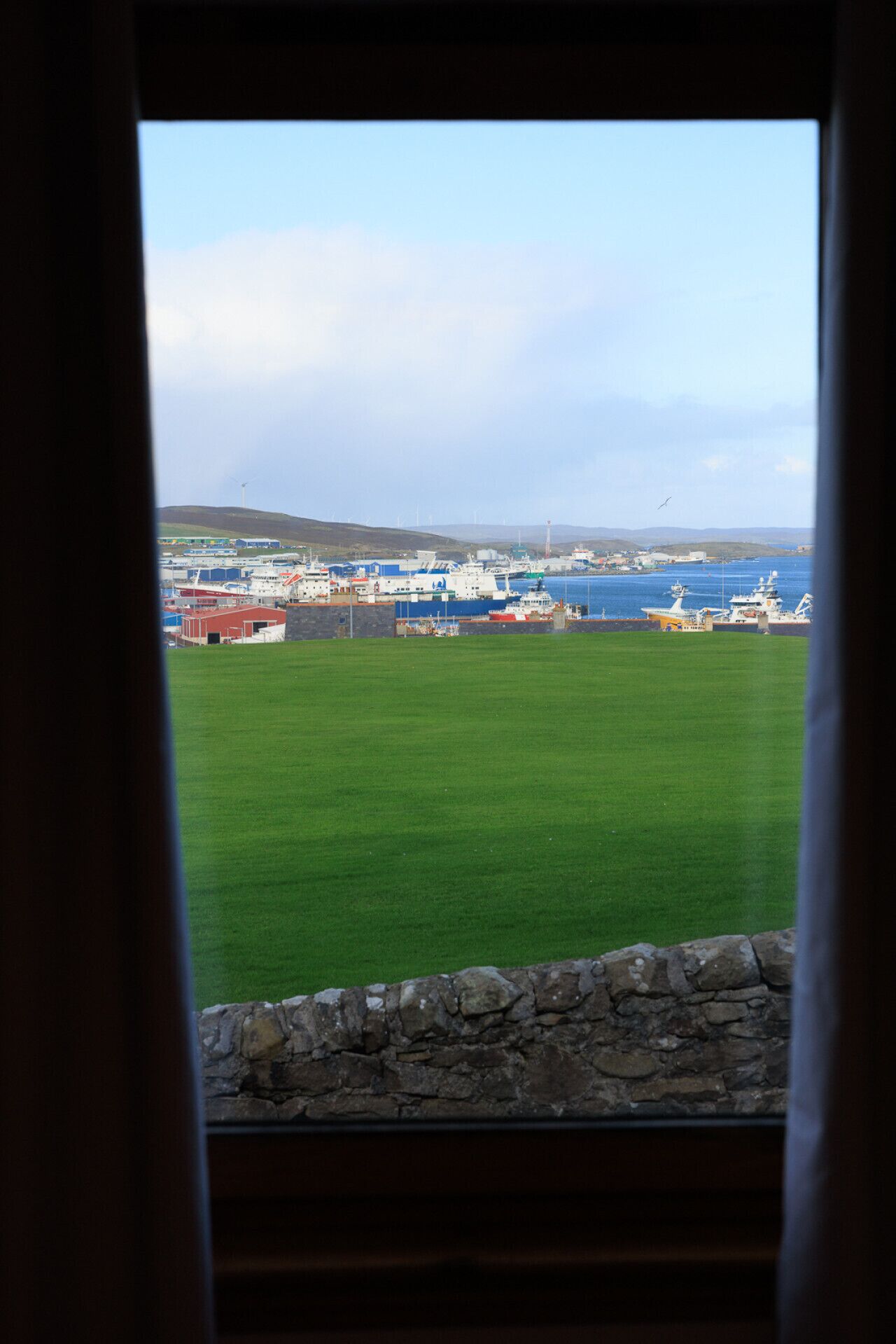 A view through a window of a grassy area with a stone wall in the foreground, leading to a marina with boats and buildings in the background.