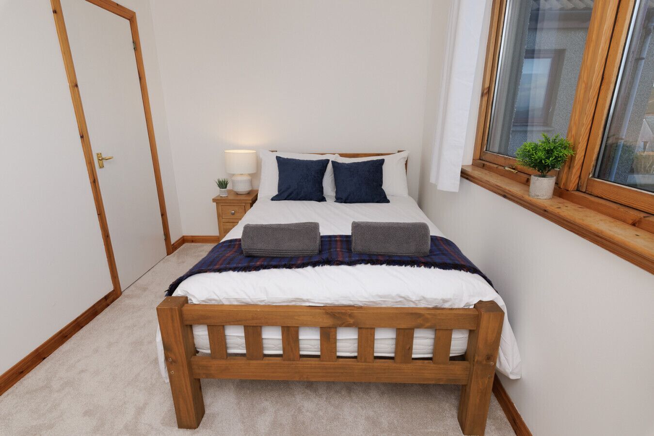 Una camera da letto con un letto in legno, biancheria bianca, un comodino con una lampada e una finestra con una pianta.