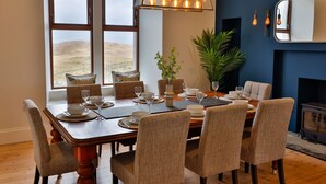 A dining area with a wooden table set for six, surrounded by wicker chairs. There is a large window with a view of a landscape, a hanging light fixture, and a potted plant on the table.