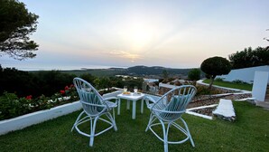 Un espace extérieur aménagé avec deux chaises et une table, offrant une vue panoramique sur un paysage pittoresque composé de collines et d’une étendue d’eau.