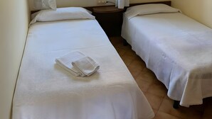 Une chambre avec deux lits, une fenêtre avec des rideaux, une étagère avec un récipient et un luminaire fixé au mur.