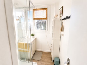 Une salle de bain avec une douche en verre, une petite fenêtre avec des stores, un meuble blanc et une poubelle bleue.